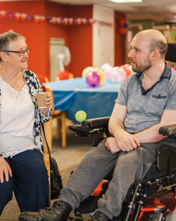 An elderly lady sat next to a man in an electric wheelchair in a community hall decorated with balloons and party decorations.